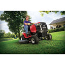 Man operating a red riding lawn mower in a grassy area with trees and a house in the background.
